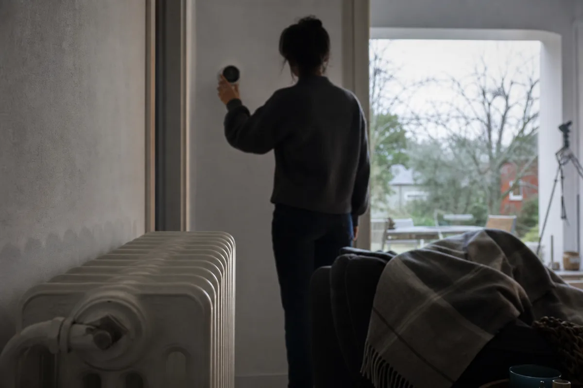 Salon d&rsquo;une maison individuelle un samedi d&rsquo;hiver, radiateur froid au premier plan, silhouette de dos vérifiant un thermostat mural, lumière douce de fin de journée